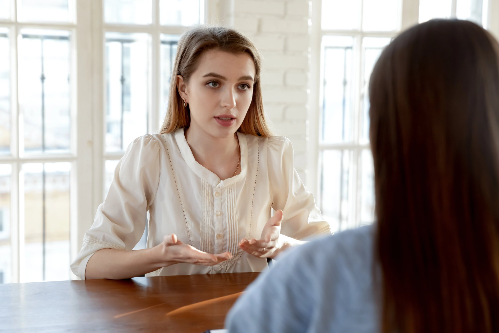 A woman with long hair is sitting at a table, gesturing while engaged in conversation with another person whose back is to the camera. They are in a bright room with large windows in the background.