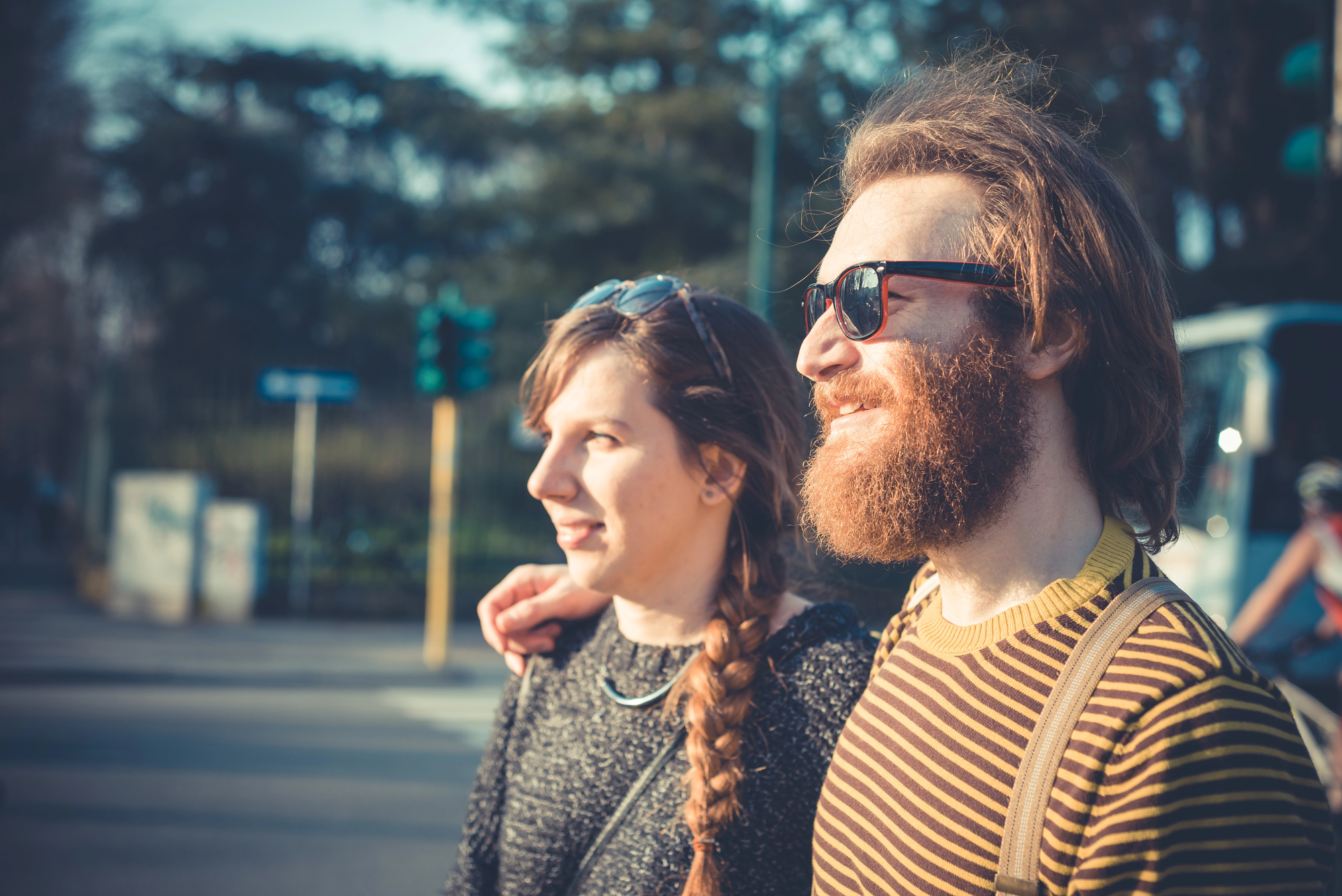 A man and woman walking outdoors on a sunny day. The woman has a braid and wears a sweater, while the man has a beard, sunglasses, and a striped shirt. They are smiling and the man has his arm around her shoulder. Trees and a street are in the background.