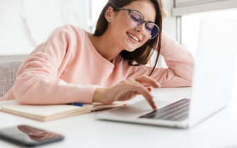 A woman wearing glasses and a pink sweater smiles while using a laptop. She is seated at a desk with a notebook and smartphone nearby. Natural light illuminates the space from a nearby window.