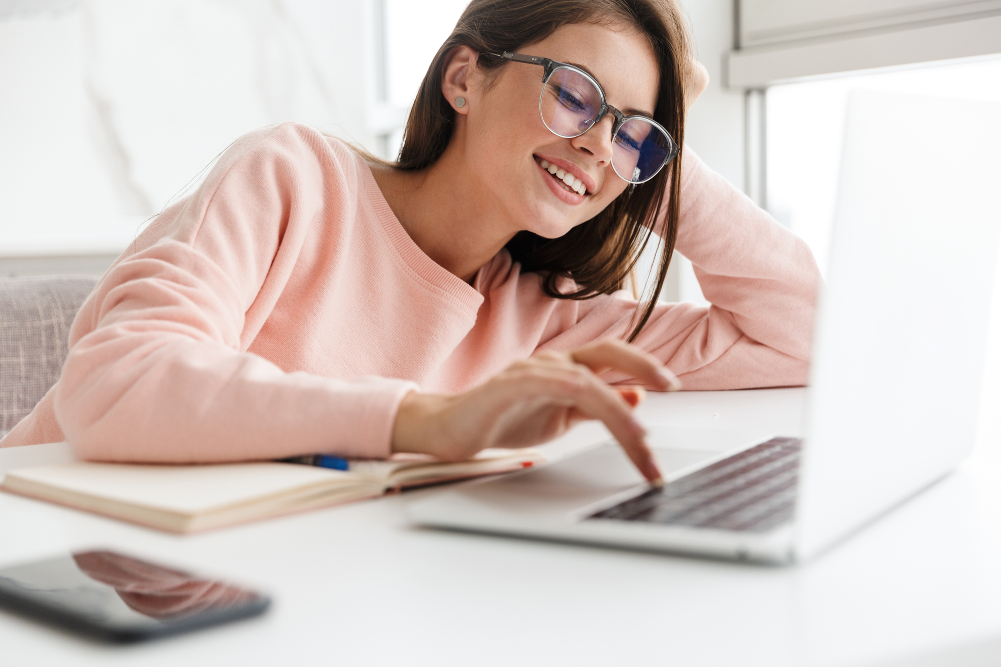 A woman wearing glasses and a pink sweater smiles while using a laptop. She is seated at a desk with a notebook and smartphone nearby. Natural light illuminates the space from a nearby window.