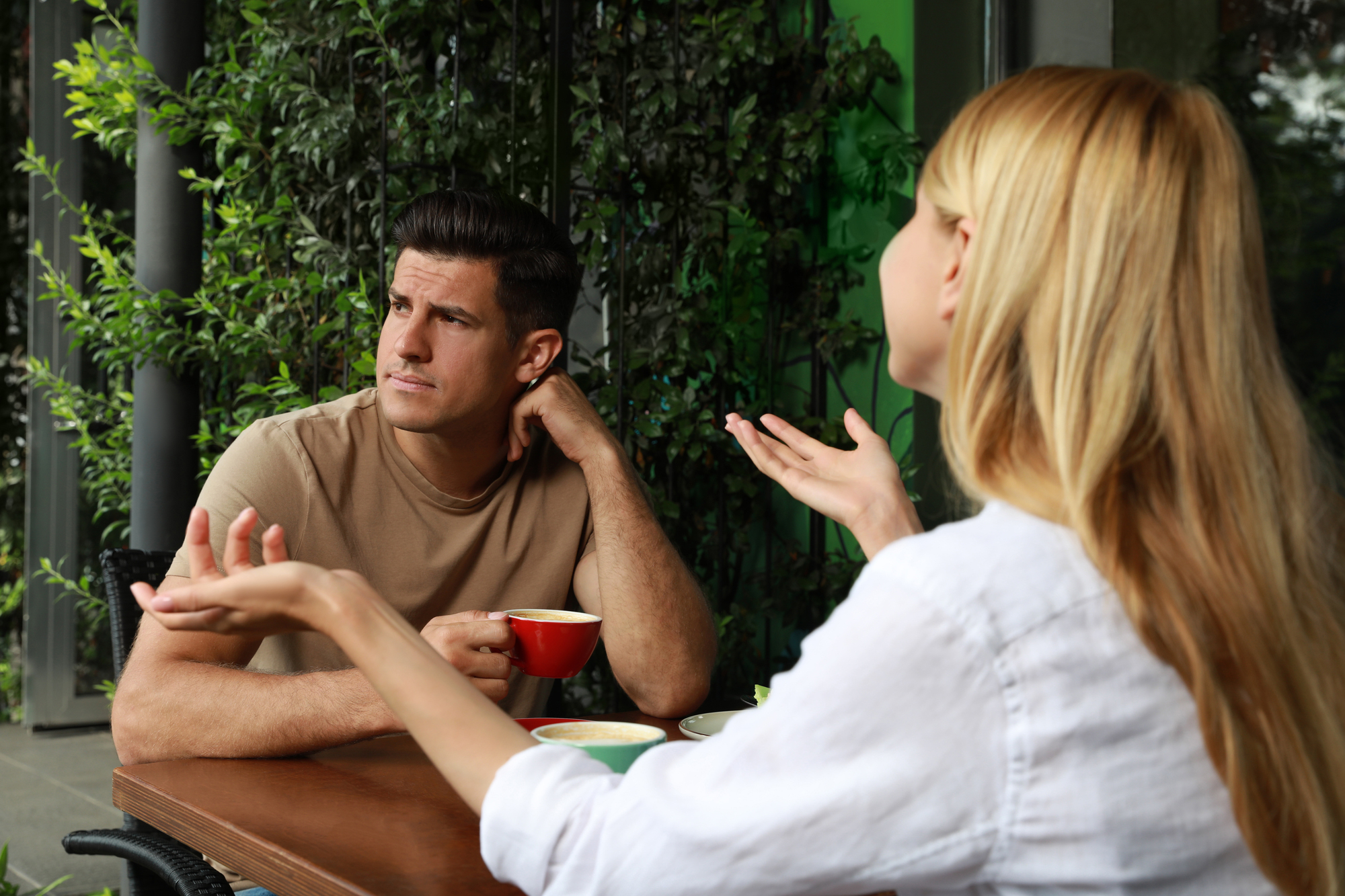 A man and woman sit at an outdoor café table. The man, holding a red coffee cup, looks away thoughtfully. The woman, with her back to the camera, gestures with her hands as if explaining something. Greenery surrounds the patio.