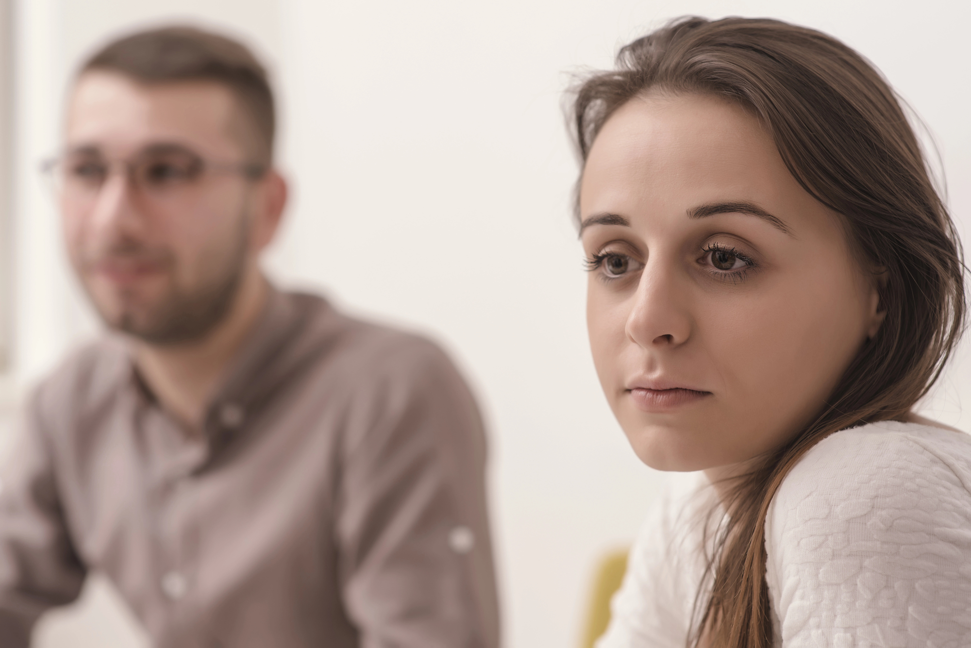 A woman with long brown hair looks pensive while sitting at a table. A man with glasses is seated beside her, slightly out of focus. The setting appears to be a neutral-toned room.