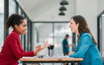 Two women sit at a table in a modern office, engaged in a lively conversation. One wears a red blazer and gestures expressively, while the other listens attentively, dressed in a blue blazer. A blurred figure and office interior are in the background.