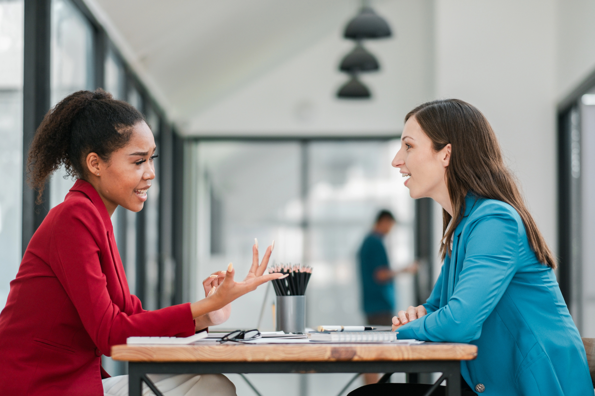 Two women sit at a table in a modern office, engaged in a lively conversation. One wears a red blazer and gestures expressively, while the other listens attentively, dressed in a blue blazer. A blurred figure and office interior are in the background.