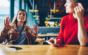 Two women sitting at a wooden table in a cozy cafe. One woman is animatedly talking with her hands raised, while the other listens attentively, holding her phone. Warm lighting and modern decor create a welcoming atmosphere.