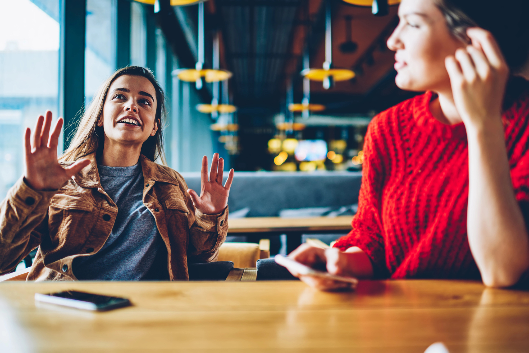 Two women sitting at a wooden table in a cozy cafe. One woman is animatedly talking with her hands raised, while the other listens attentively, holding her phone. Warm lighting and modern decor create a welcoming atmosphere.