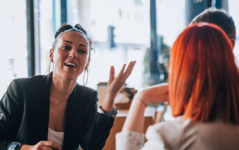 A woman with hoop earrings and a bun gestures while talking to a person with red hair in a brightly lit cafe. They are seated at a table, engaged in conversation.