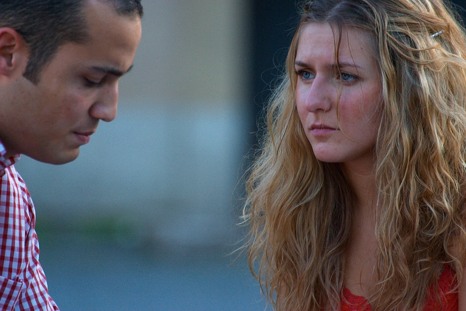 A man and a woman are sitting closely together outdoors. The man is looking down, his expression serious, while the woman, with long blonde hair, looks intently ahead, appearing deep in thought or concerned. Both are wearing casual clothing.