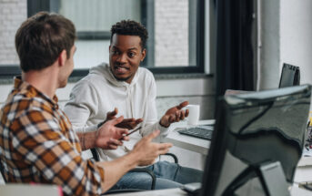 Two men are in an office setting, engaged in a lively discussion. One wears a white hoodie while the other is in a plaid shirt. They are seated in front of computer monitors, gesturing expressively as they talk.