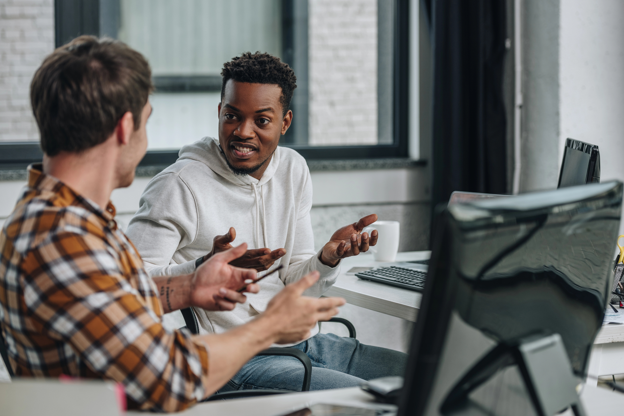 Two men are in an office setting, engaged in a lively discussion. One wears a white hoodie while the other is in a plaid shirt. They are seated in front of computer monitors, gesturing expressively as they talk.