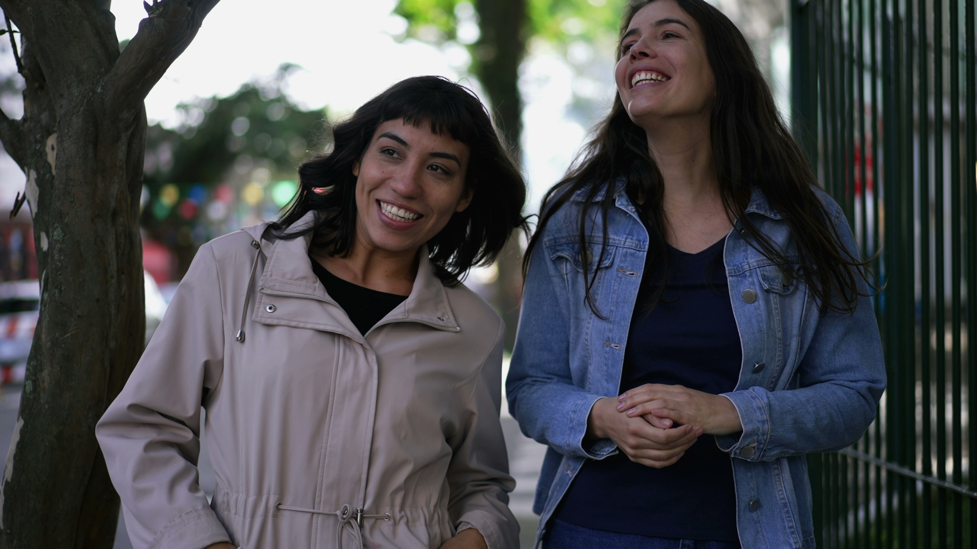 Two women are walking and laughing together outdoors. One wears a beige jacket and the other a denim jacket. They are on a tree-lined street with colorful flags in the background, enjoying a sunny day.