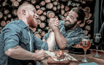 Two men with beards are arm wrestling at a wooden table in a rustic setting, surrounded by logs. They both appear enthusiastic and competitive. A plate with food and two glasses of beer are on the table.