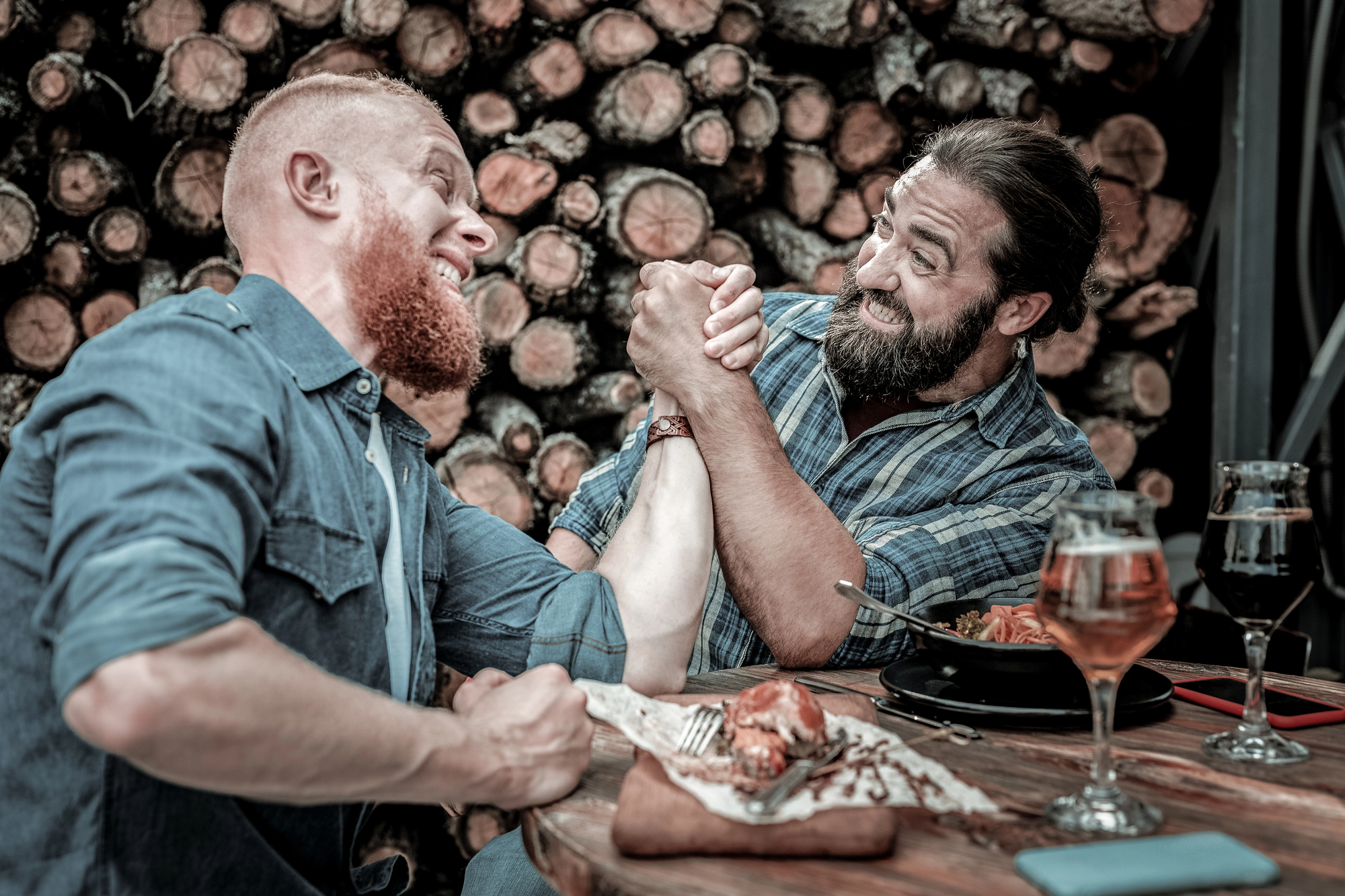 Two men with beards are arm wrestling at a wooden table in a rustic setting, surrounded by logs. They both appear enthusiastic and competitive. A plate with food and two glasses of beer are on the table.