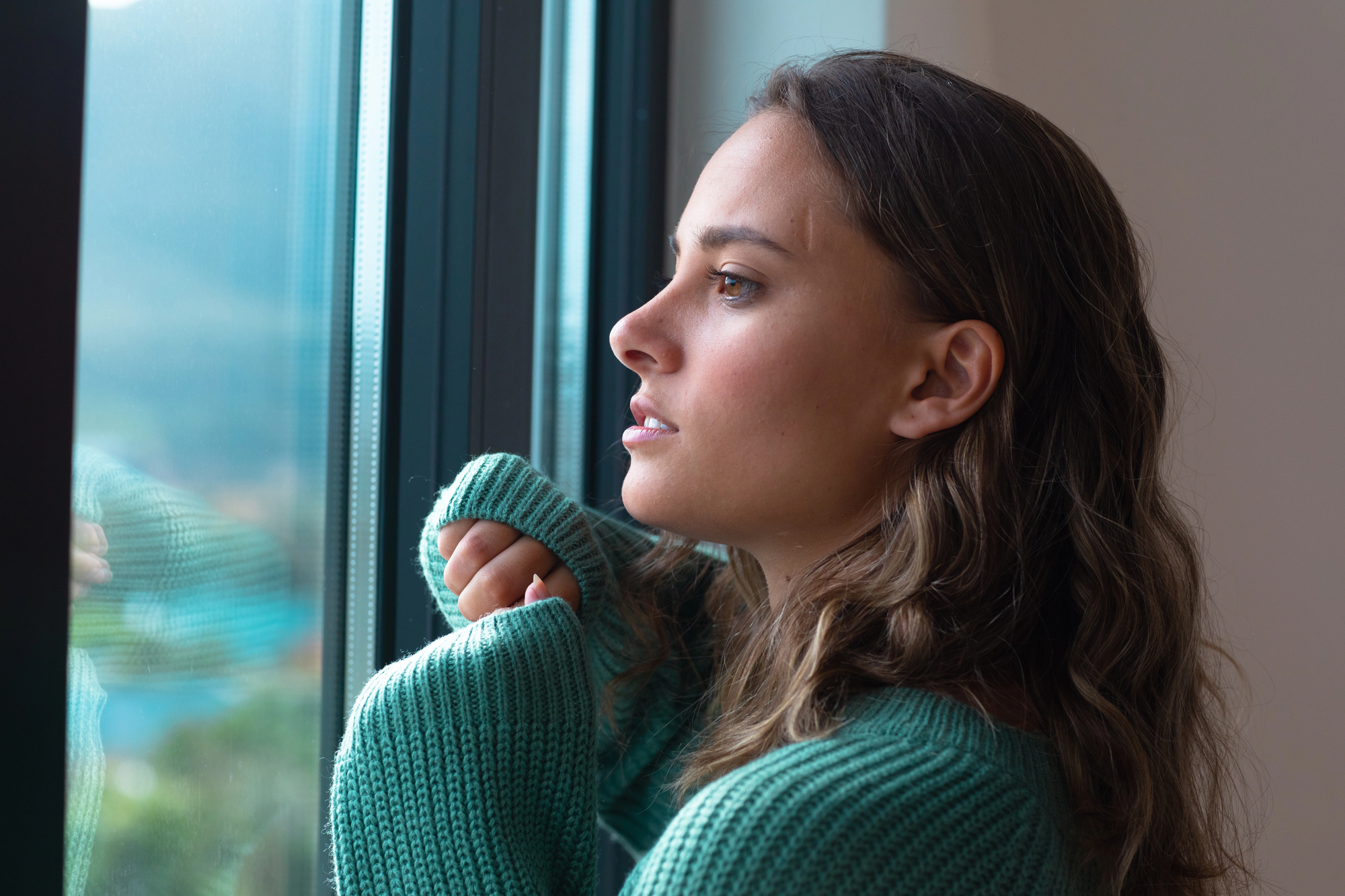 A person with long hair gazes thoughtfully out a window, wearing a teal sweater. The background shows a blurred view of the outdoors, conveying a sense of calm and introspection.