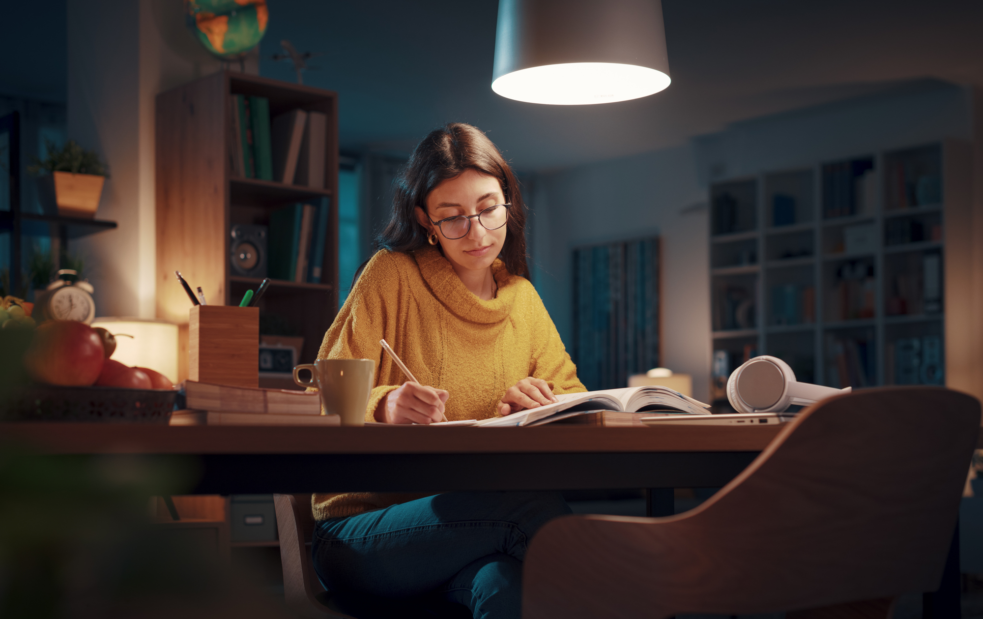 A woman wearing glasses and a yellow sweater sits at a desk writing in a notebook. The room is dimly lit, with a lamp overhead. Books, a globe, and a cup are on the desk. Shelves and a clock are visible in the background.