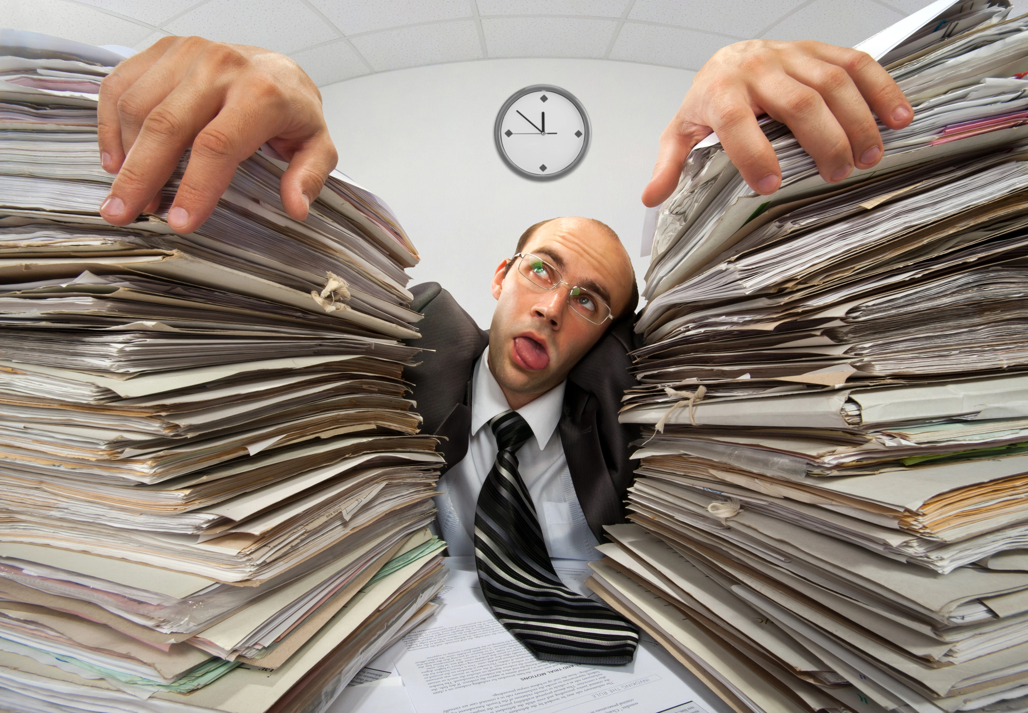 A man in a suit appears overwhelmed as he sits between two large stacks of paperwork. He is making a humorous expression with his eyes crossed and tongue out. A clock showing 3:55 is on the wall behind him.