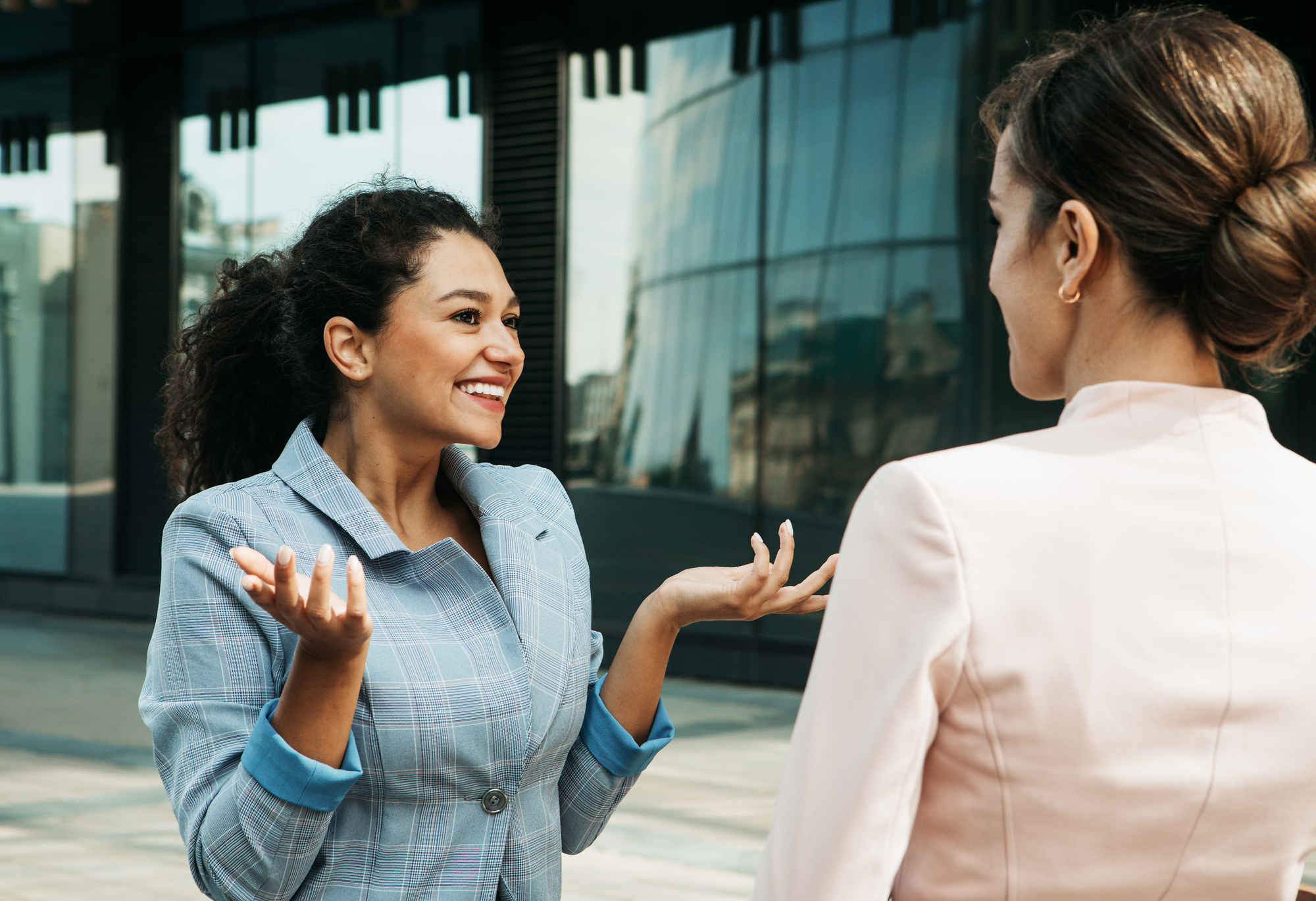 Two women are having an animated conversation outside a modern glass building. The woman on the left gestures expressively with a smile, while the woman on the right listens attentively, her back facing the camera.