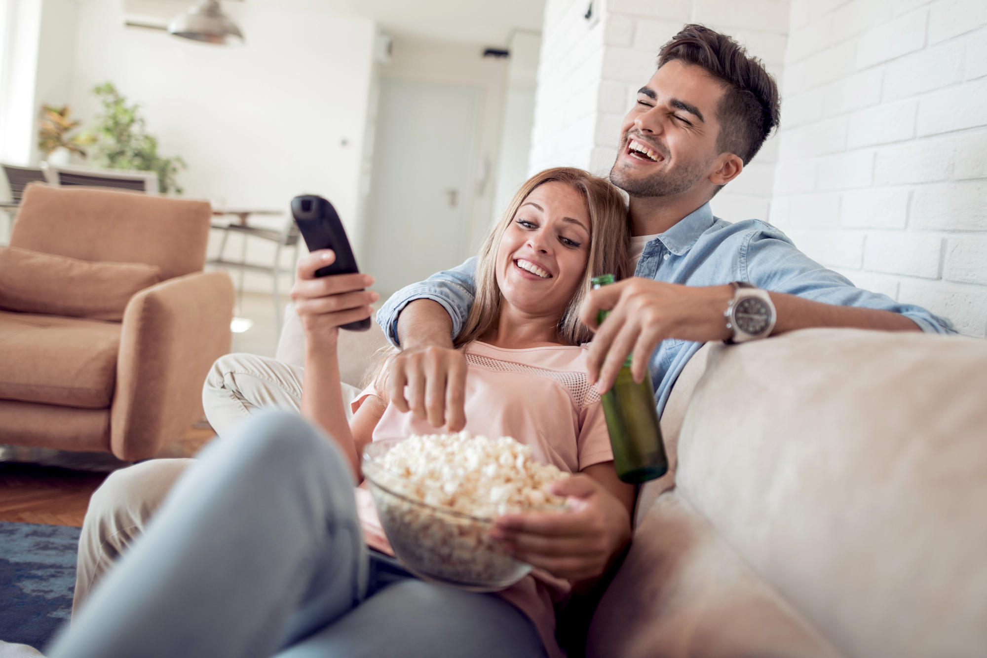 A couple sits on a sofa, smiling and relaxed. The woman holds a remote control and a bowl of popcorn, while the man, holding a beer, leans back. The room has a cozy atmosphere with modern decor.