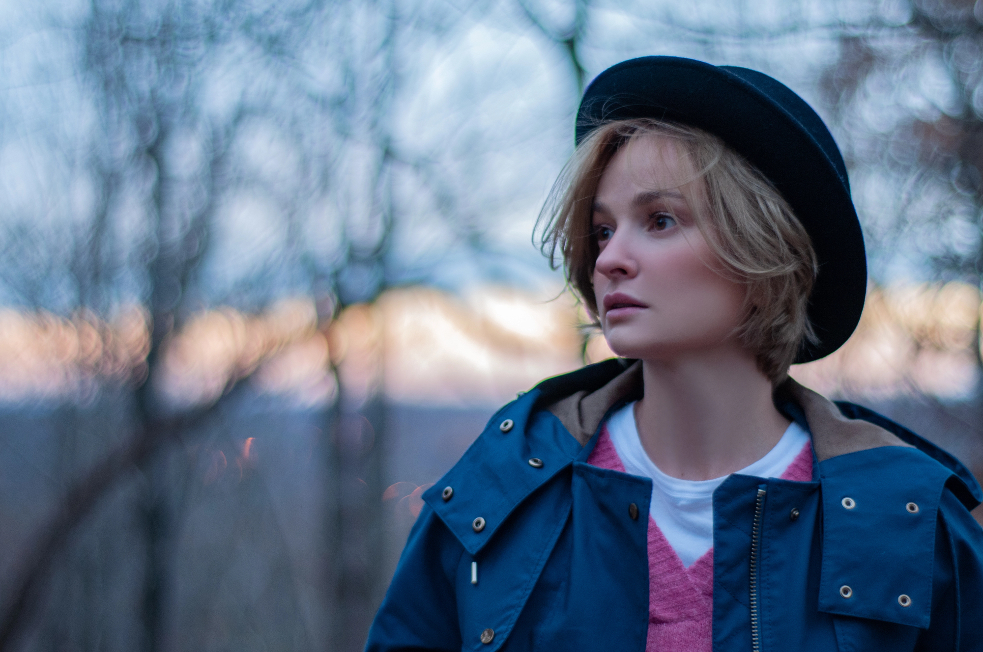 A person with short blonde hair wears a dark hat and a blue jacket over a pink sweater, standing outdoors with bare trees in the background. The sky is softly lit by a setting or rising sun.