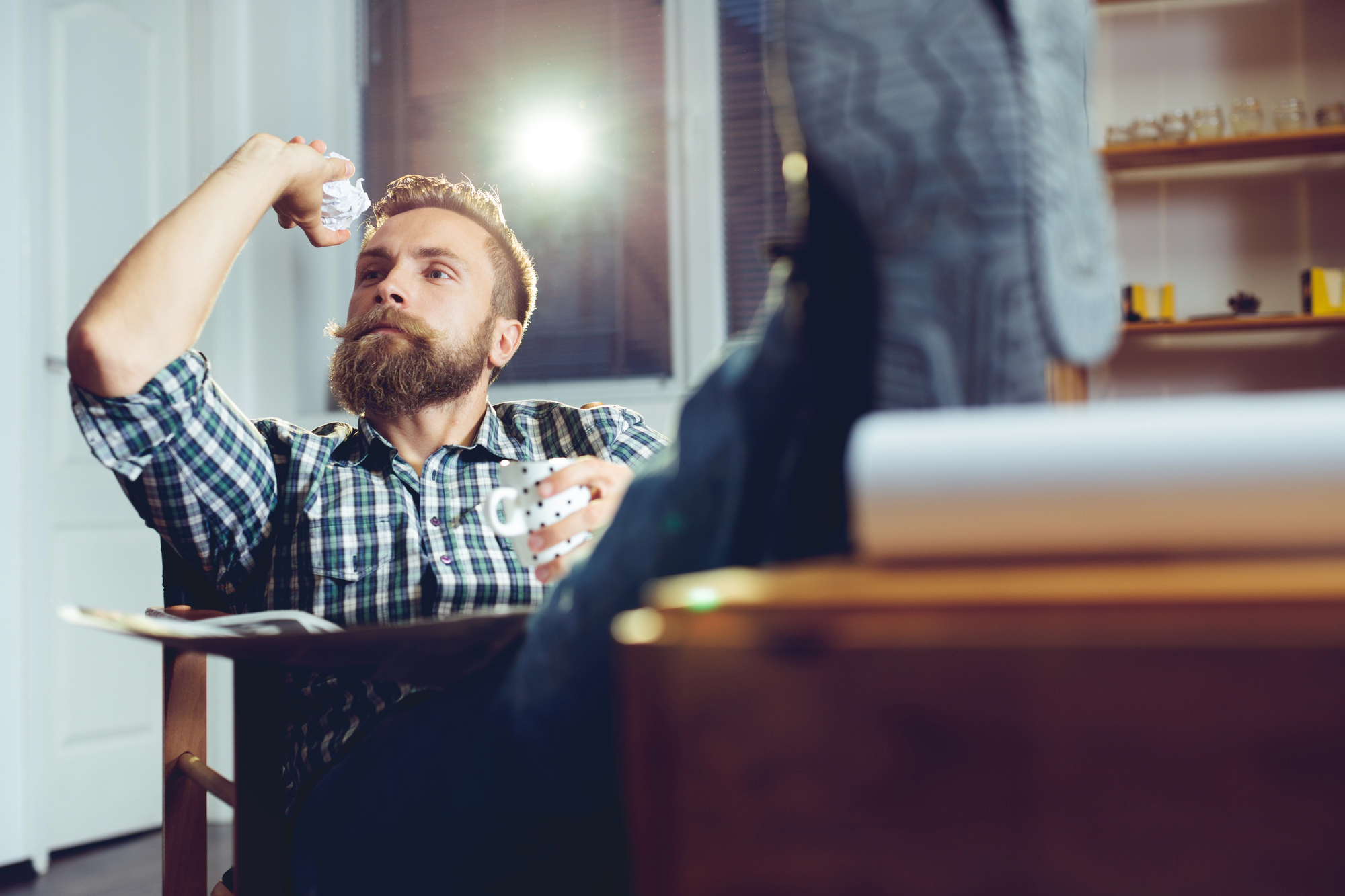 A bearded man in a plaid shirt sits casually with his feet up on a desk. He holds a mug in one hand and has a pen in his mouth, while tossing a crumpled paper ball into the air. The scene is relaxed and informal.