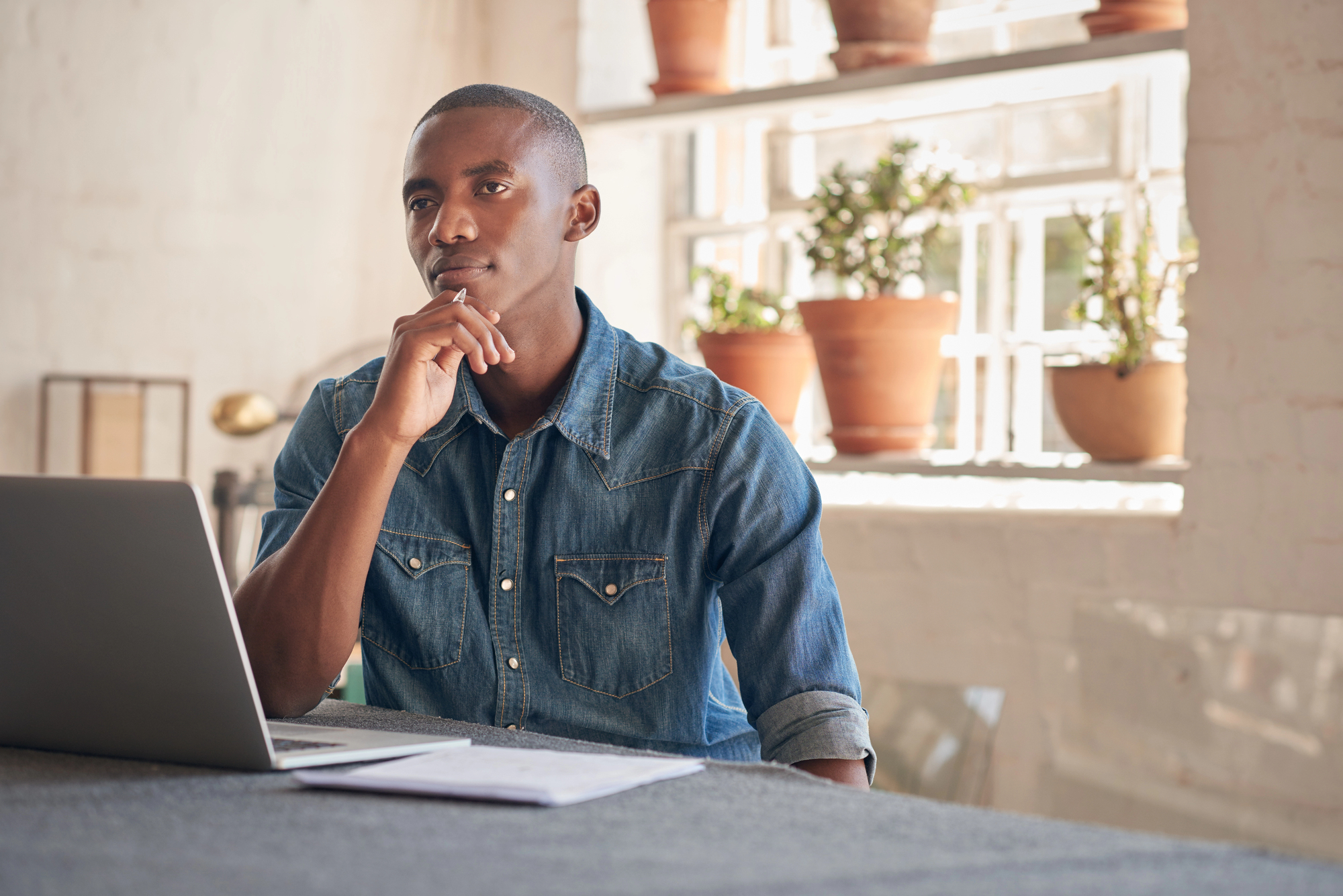 A man in a denim shirt sits at a table, thoughtfully looking at a laptop. Sunlight streams through a window with potted plants on the sill in the background. Papers lie on the table nearby.