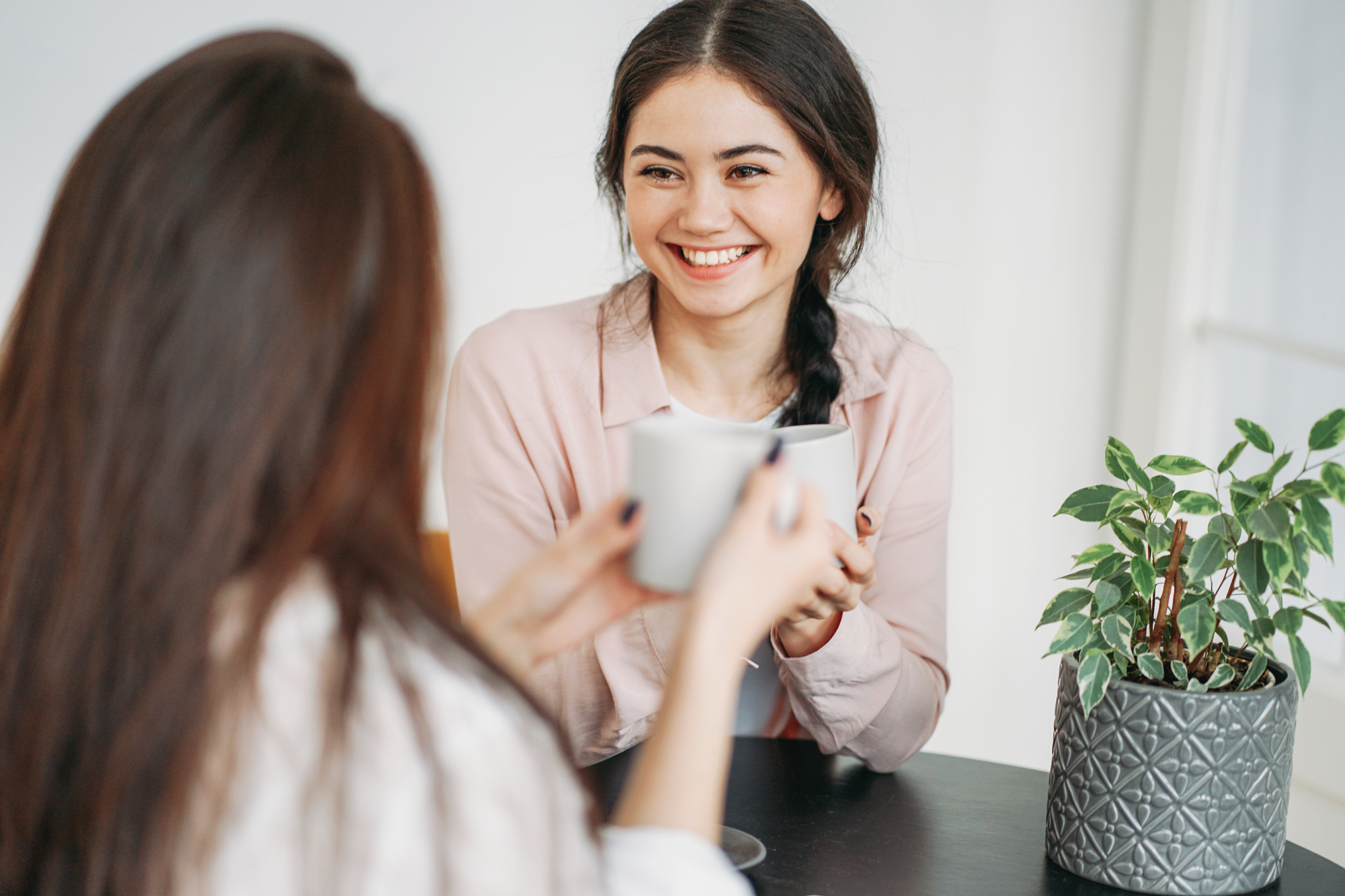 Two women sit at a table enjoying coffee together. One woman faces the camera, smiling and holding a mug, while the other has her back turned. A small potted plant is on the table in the foreground.