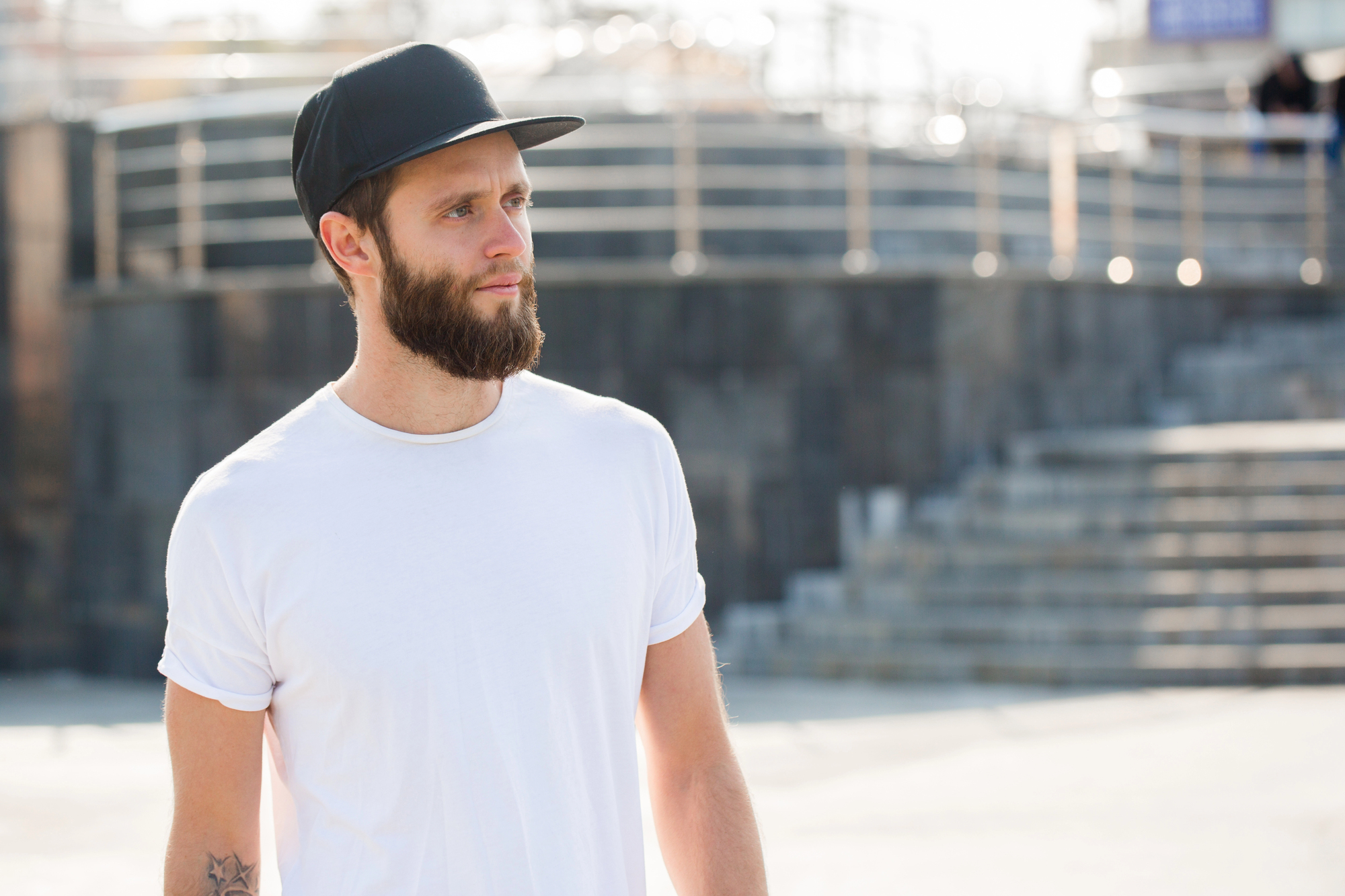 A man with a beard wearing a black cap and white t-shirt stands outdoors. He is looking to the side, and the background features blurred stairs and a railing. The image is taken in bright daylight.