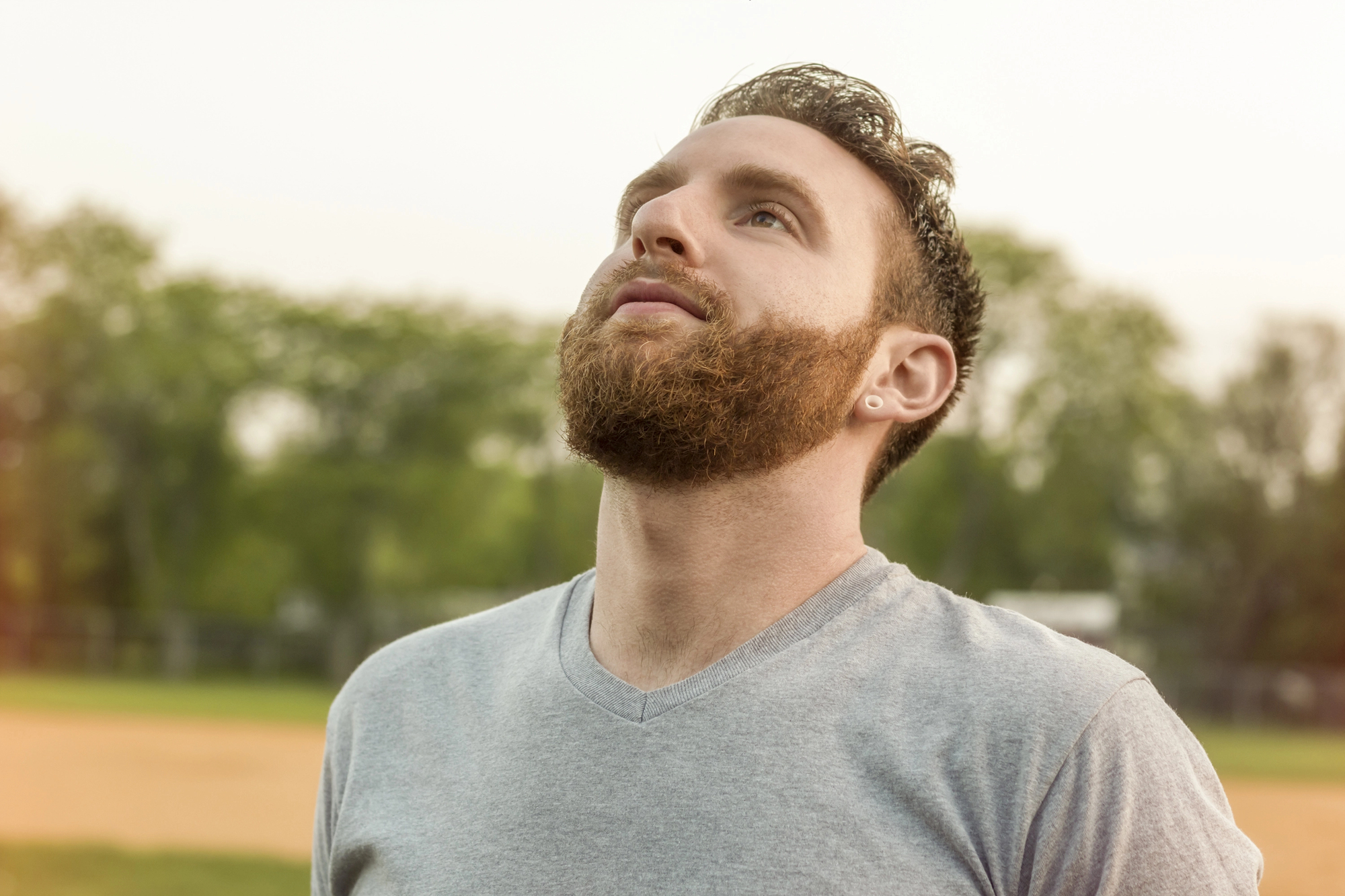 A man with a beard is standing outdoors on a sunny day, looking upwards with a thoughtful expression. He is wearing a gray t-shirt, and there are trees and a grassy field in the background.