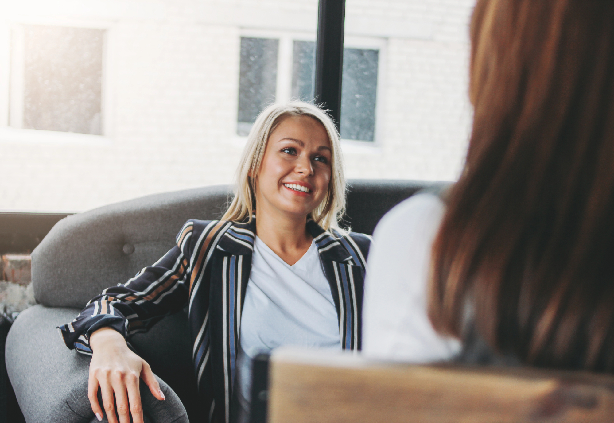 A woman with blonde hair, wearing a striped blazer and white shirt, sits on a gray couch, smiling while talking to another person whose back is facing the camera. They are in a bright room with large windows.