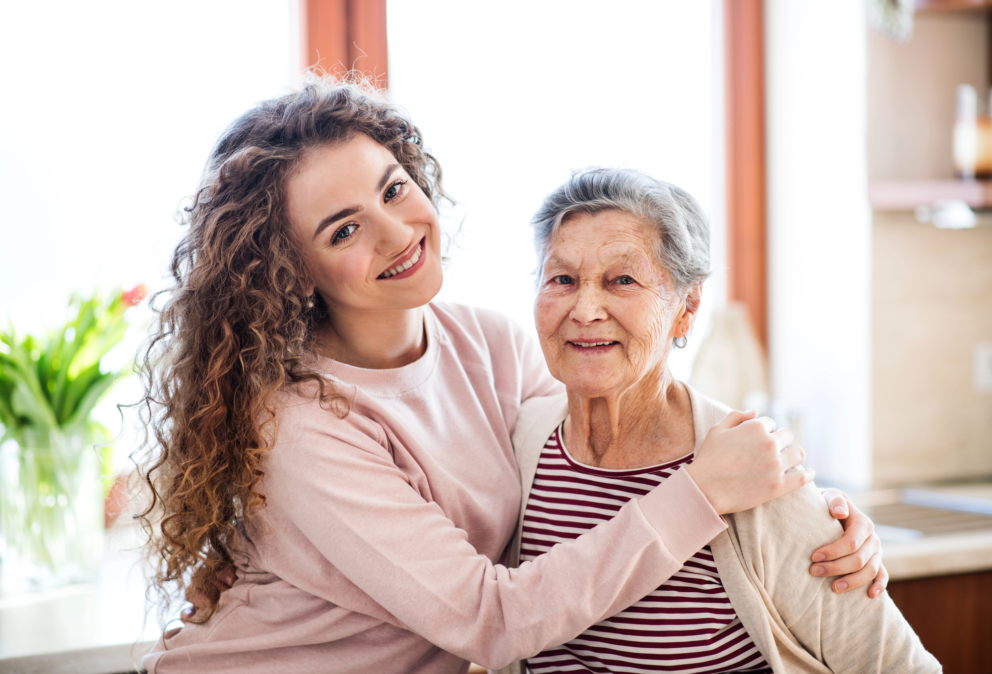 A young woman with curly hair embraces an elderly woman with short gray hair in a warmly lit room. They both smile at the camera, with potted plants visible in the background.