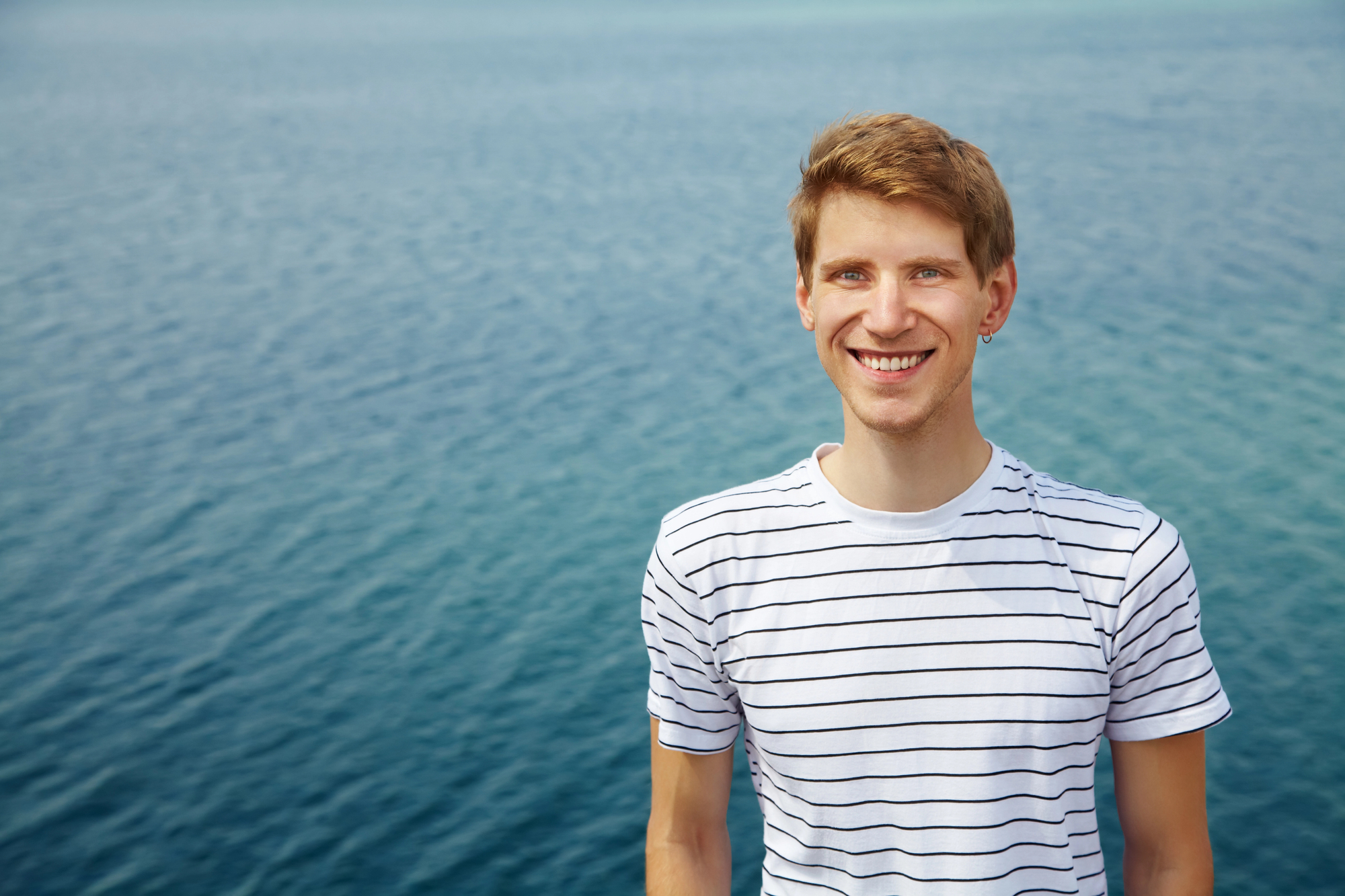 A young man in a striped shirt smiles at the camera, standing in front of a vast blue ocean under a clear sky.