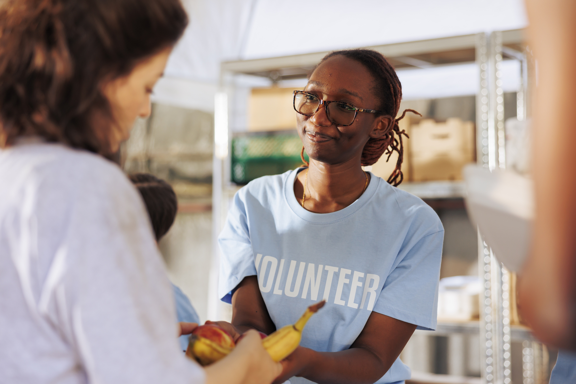 A volunteer wearing a blue shirt helps distribute bananas to a woman at a food bank. They are under a tent with shelves in the background containing various food items. The setting appears to be a community service event.