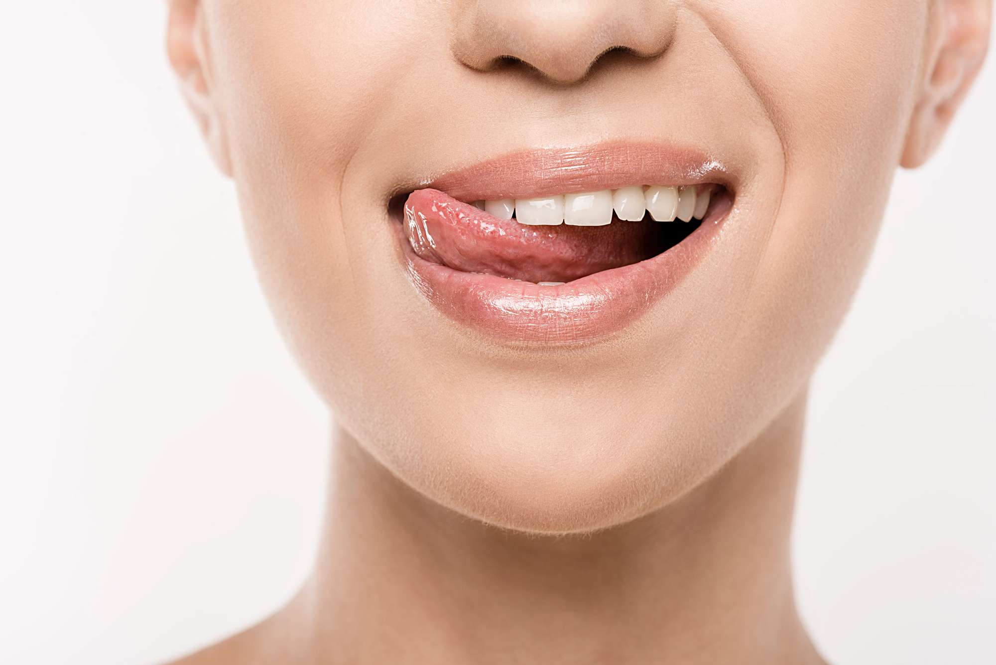 Close-up of a person's face with smooth skin, showing their mouth and part of their nose. Their lips are glossy as they smile while playfully sticking out their tongue to lick the upper lip.