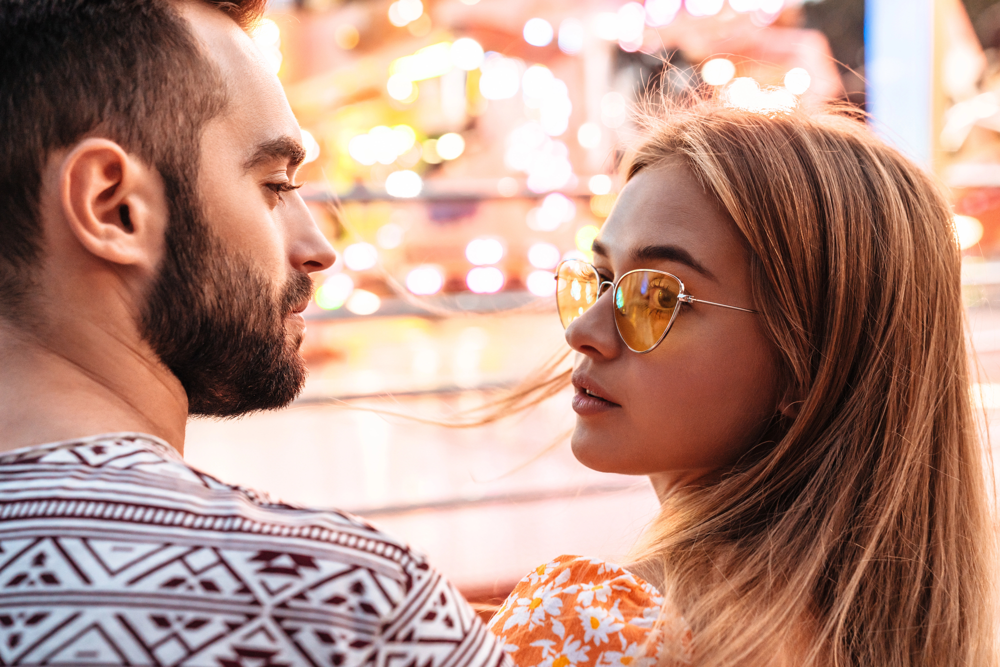 A man and woman stand close together, facing each other. The woman, wearing sunglasses, looks at the man. Colorful, blurred lights in the background suggest a lively environment, possibly an amusement park. Both appear engaged and relaxed.