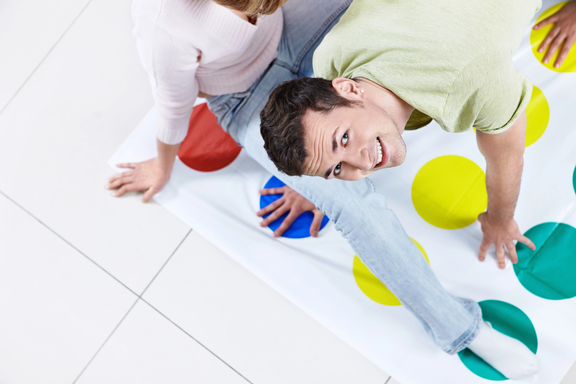 Two people are playing Twister on a tiled floor. The man in the foreground, wearing a green shirt and jeans, smiles at the camera. The woman beside him, in a light-colored sweater, is facing away. The mat is covered with colorful dots.