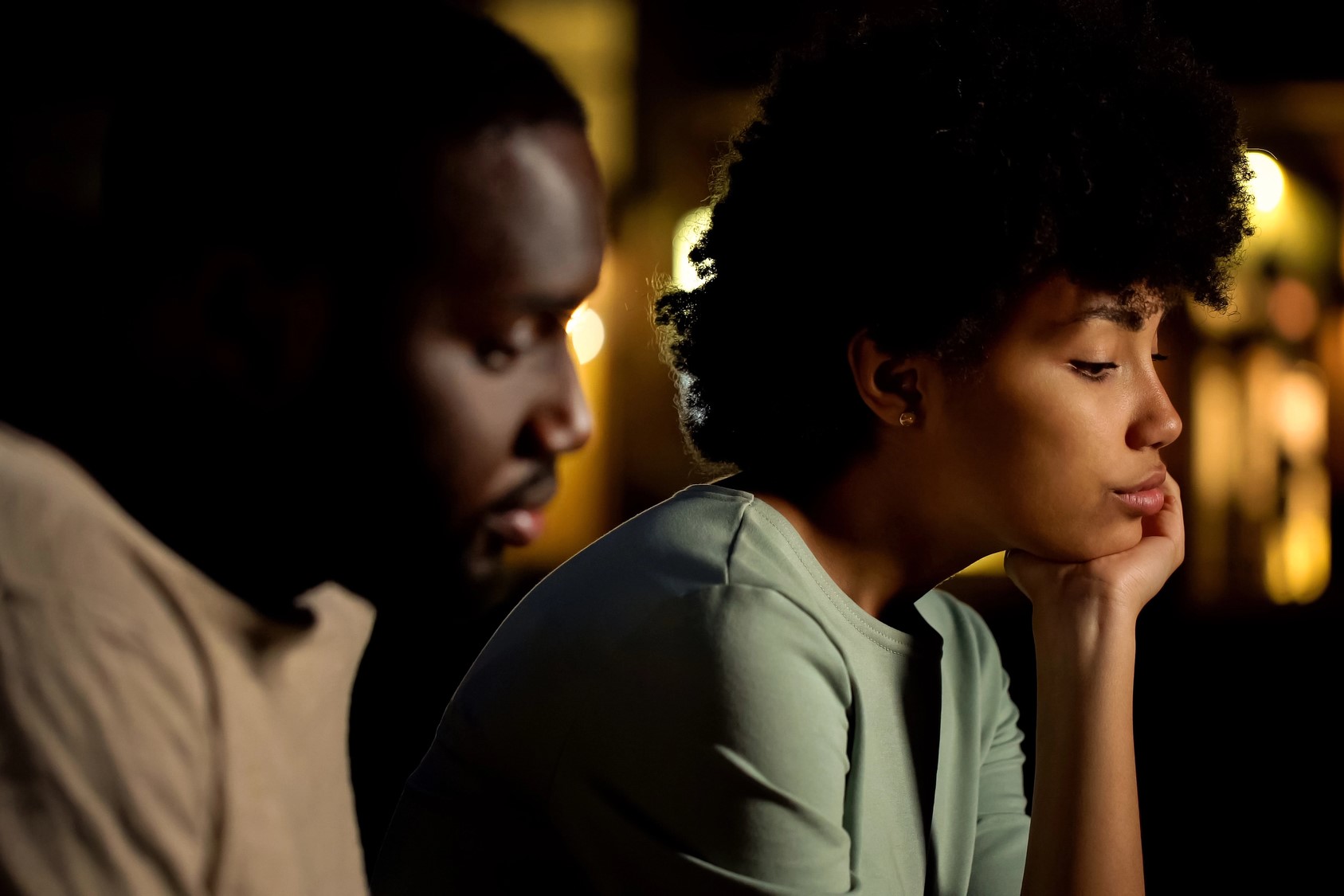 A man and woman sit close together in a dimly lit room. The woman is resting her chin on her hand, looking thoughtful. The man appears to be in contemplation. Soft, blurred lights can be seen in the background.