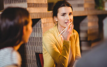 A woman in a yellow sweater holds a pencil and appears thoughtful, sitting at a table with two other people in a warmly lit room.