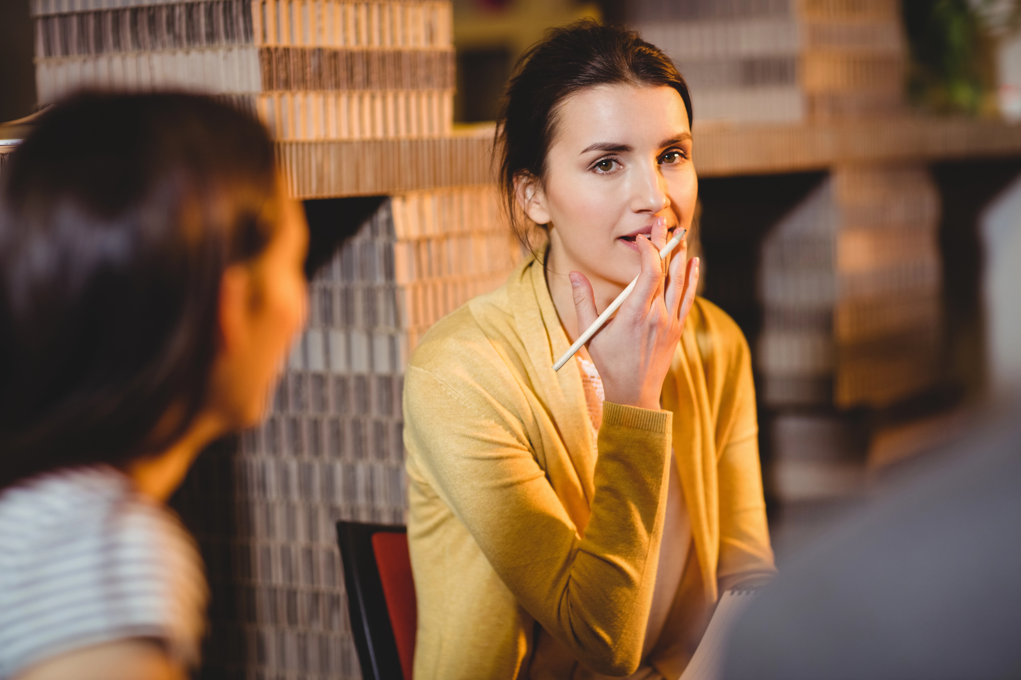 A woman in a yellow sweater holds a pencil and appears thoughtful, sitting at a table with two other people in a warmly lit room.