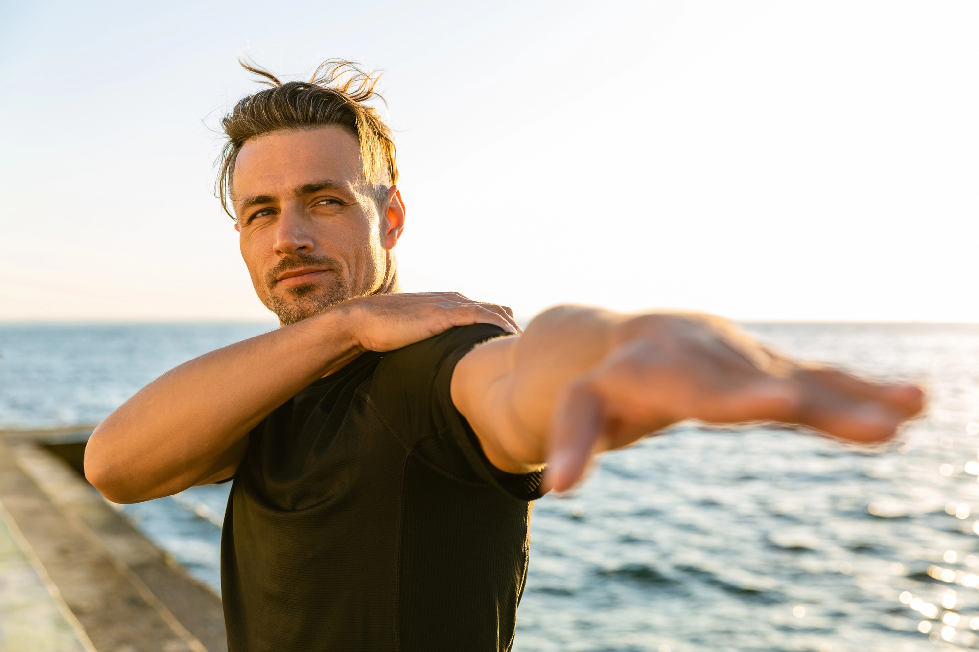 A man with short hair stretches his arm out to the side while holding his shoulder with the other hand. He is wearing a black shirt and standing by the sea during sunset, with the sun creating a warm glow in the background.