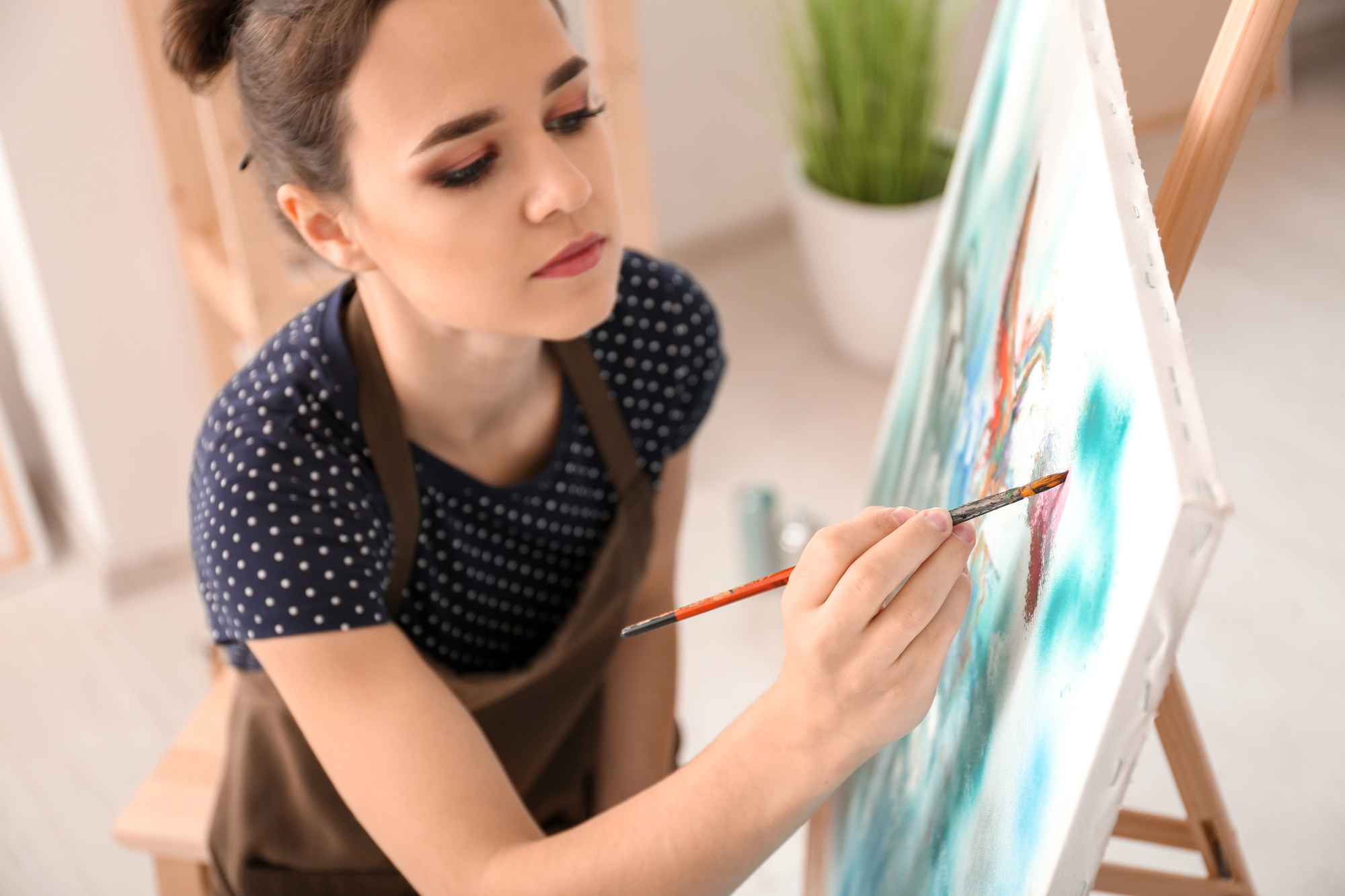 A woman in a polka dot shirt and apron is focused on painting a colorful abstract piece on a large canvas set on an easel. She is seated in a brightly lit room with a potted plant in the background.