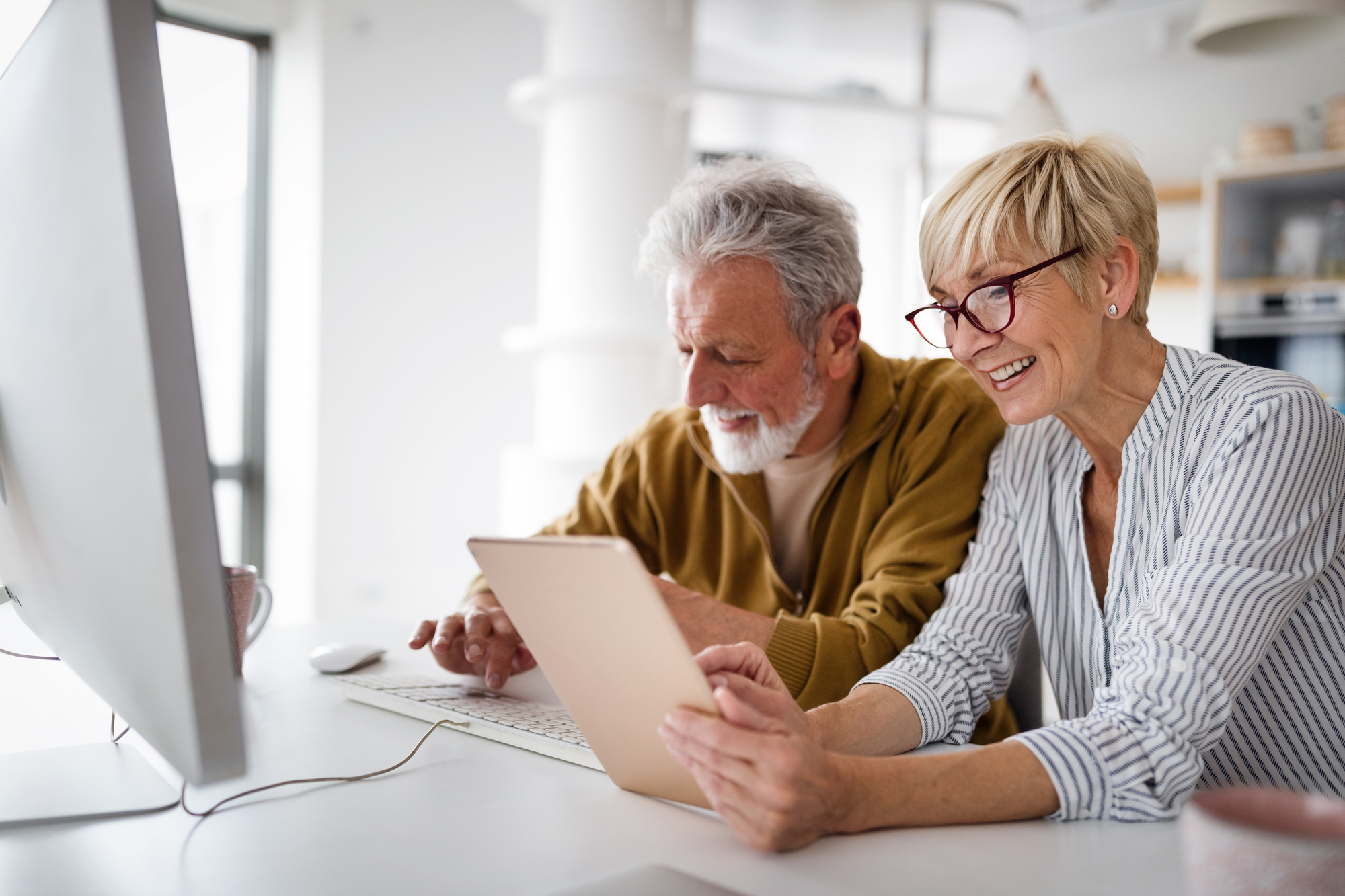 An elderly couple sits at a desk, smiling. The man types on a keyboard in front of a desktop computer, while the woman looks at a tablet. The setting appears to be a bright and modern home office.