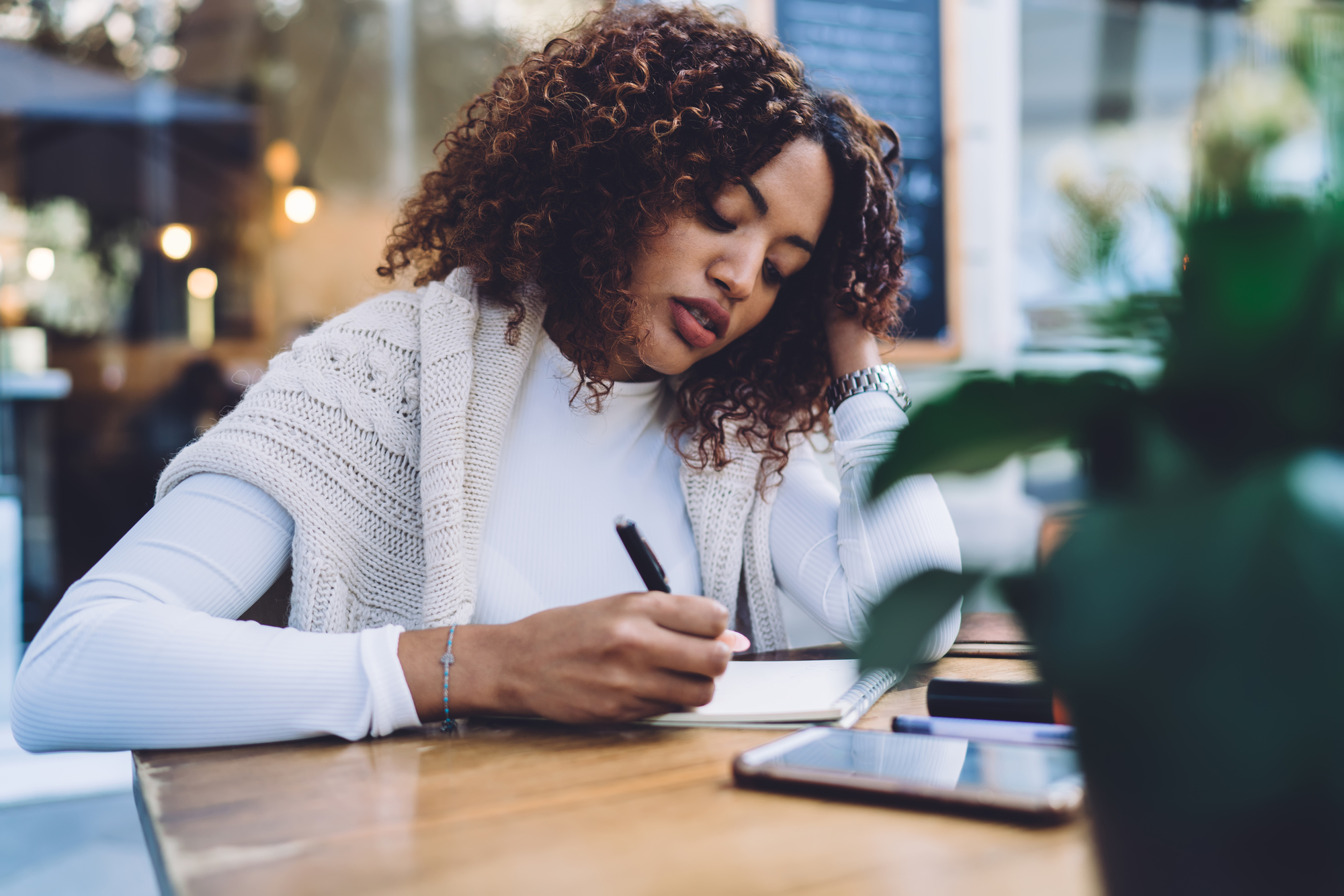 A woman with curly hair is sitting at a wooden table in a café, writing in a notebook. She is wearing a white top and a knitted cardigan. Her phone and a potted plant are on the table. There is a chalkboard menu in the background.