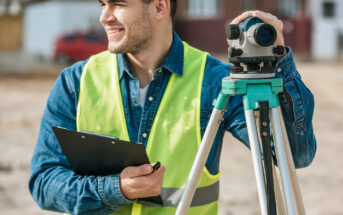 A construction worker in a yellow vest and helmet smiles while holding a clipboard next to a surveying instrument. The setting appears to be a construction site, with blurred buildings in the background.