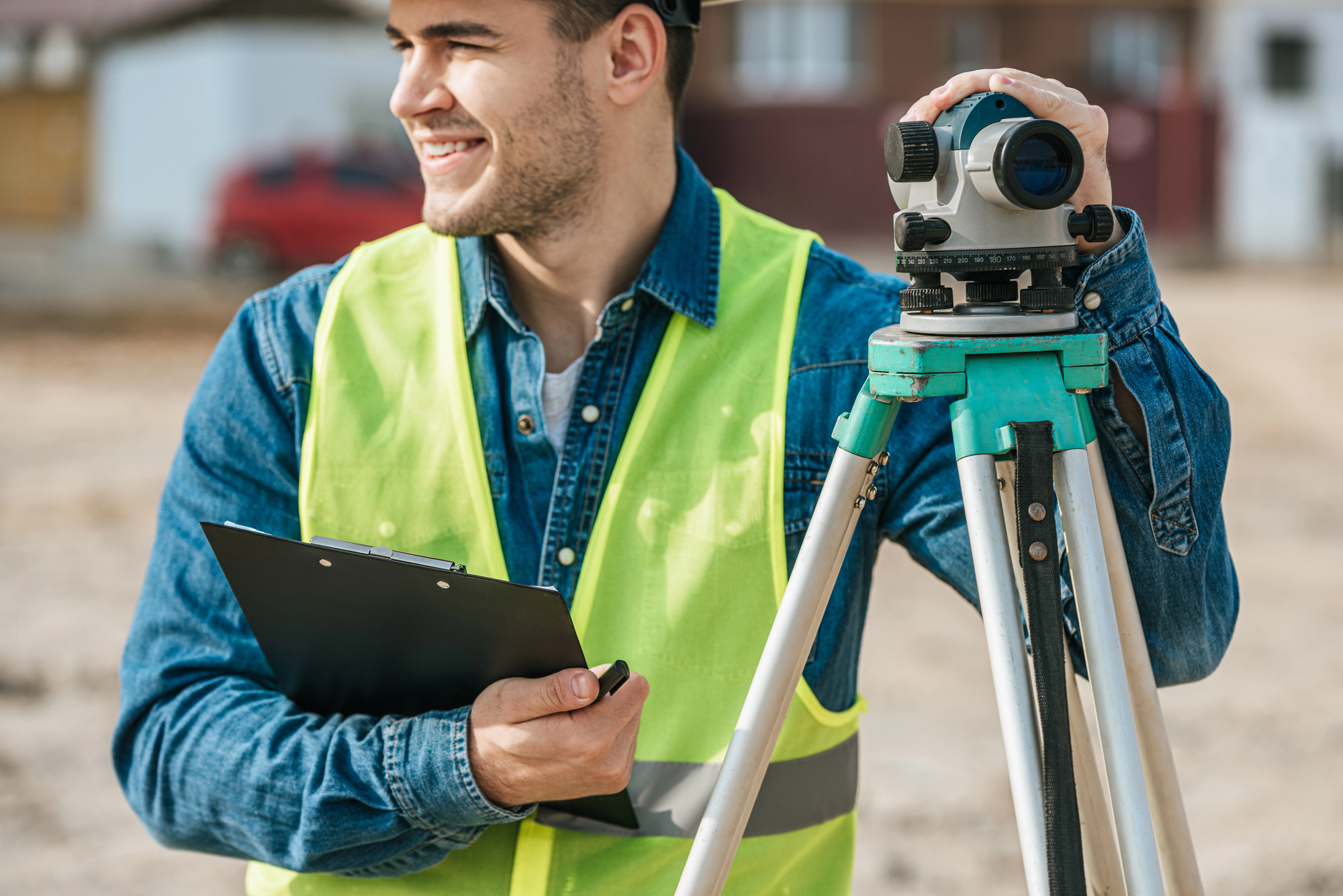 A construction worker in a yellow vest and helmet smiles while holding a clipboard next to a surveying instrument. The setting appears to be a construction site, with blurred buildings in the background.