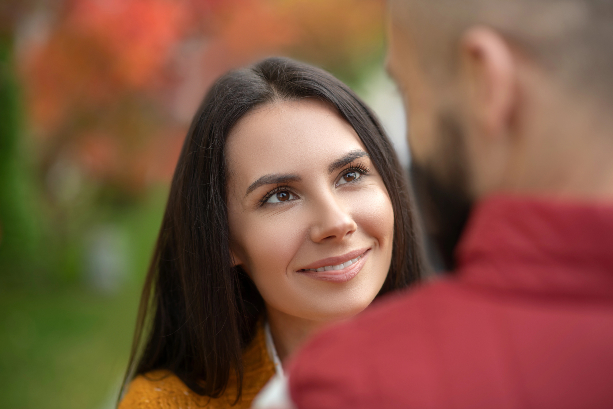 A woman with long dark hair smiles at a man in an outdoor setting with autumn-colored leaves in the background. The man is seen from behind, wearing a red jacket, while she wears a warm-toned sweater.