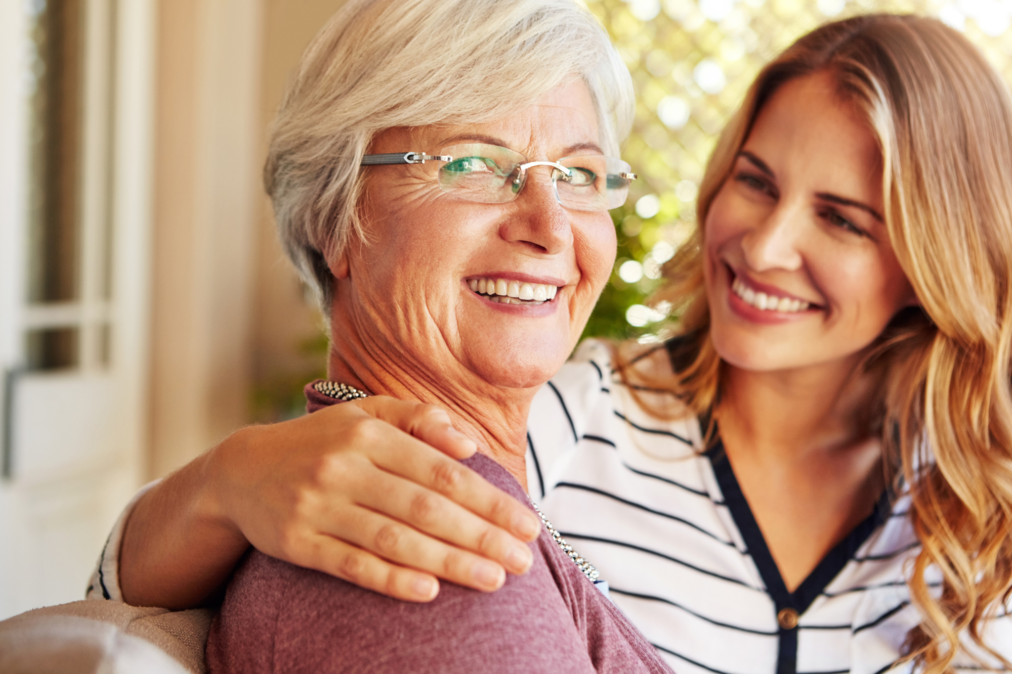 An older woman with glasses smiles broadly, while a younger woman with long hair and a striped shirt lovingly places an arm around her shoulder. Both appear happy and are indoors, with a blurred background.