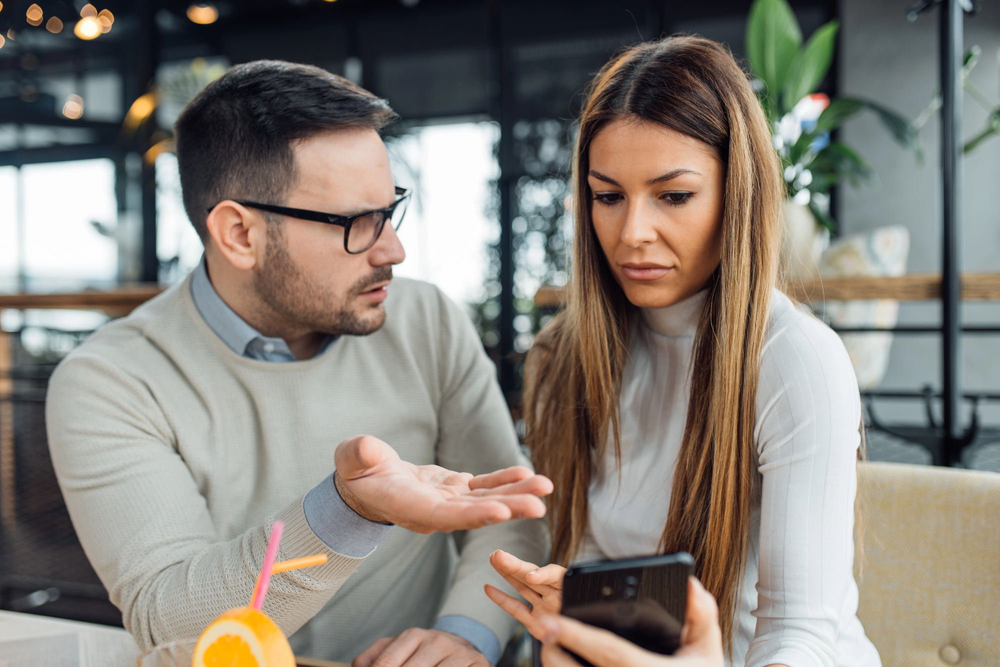 A man and woman sit in a café, engaged in a serious conversation. The man gestures with his hand, while the woman looks at a smartphone in her hand. A glass of orange juice with a straw and umbrella sits on the table in front of them.