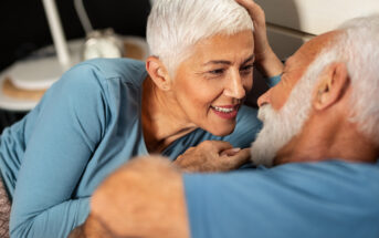 An elderly couple with white hair and wearing blue shirts gazes affectionately at each other while lying on a bed. The woman is smiling and gently touching the man's head. A clock is visible on a bedside table in the background.