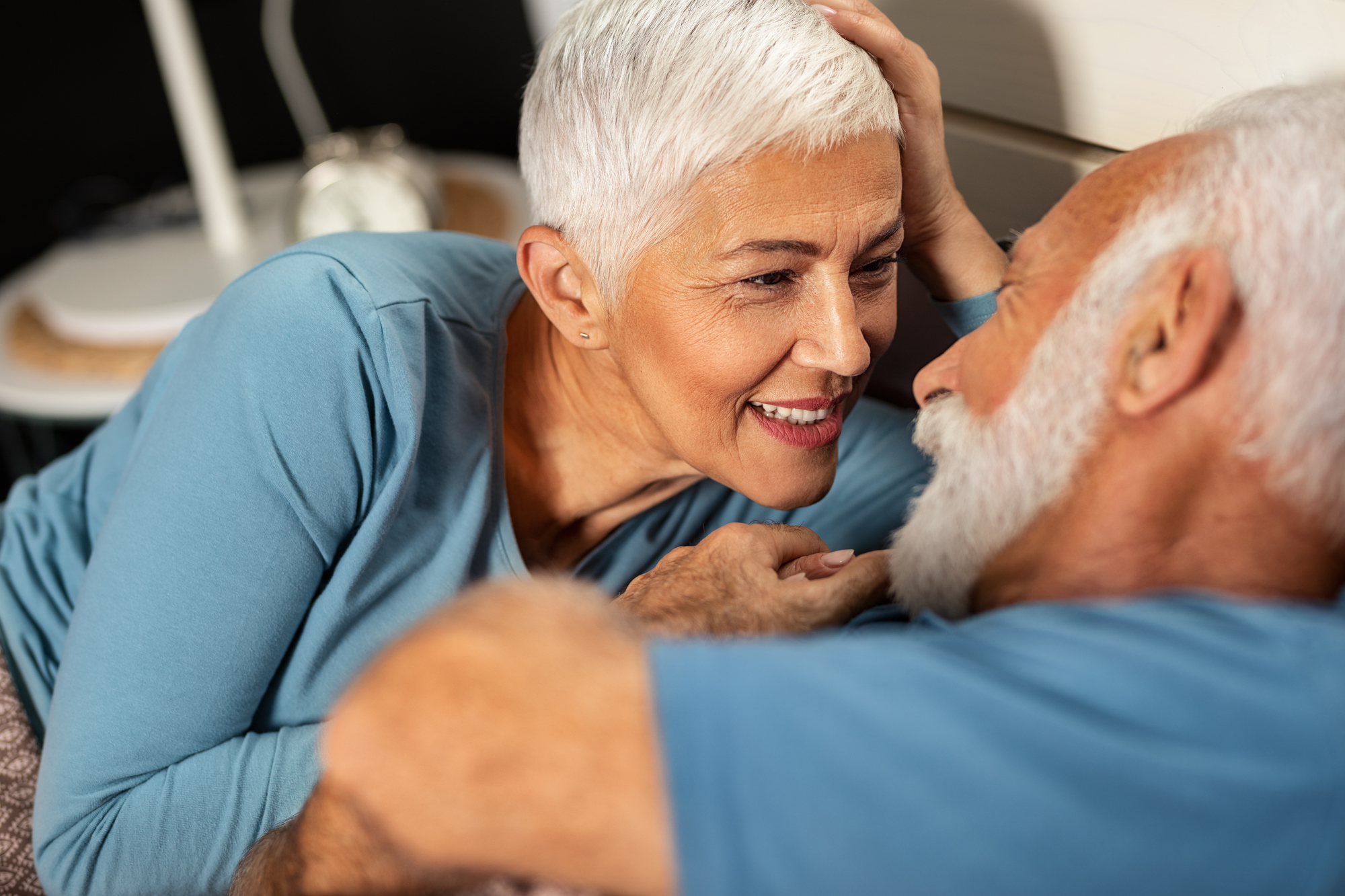 An elderly couple with white hair and wearing blue shirts gazes affectionately at each other while lying on a bed. The woman is smiling and gently touching the man's head. A clock is visible on a bedside table in the background.