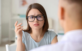 A woman with glasses and a focused expression is sitting and gesturing with her hand, looking at a man whose back is to the camera. The setting appears to be an office or meeting room.